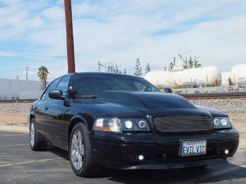 2003 Ford Crown Victoria - black sedan with a billet grille, halo projector lights and a California EVIL VIC plate parked near industrial rail tanks.