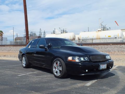 2003 Ford Crown Victoria - Front three-quarter view of a lowered black sedan on chrome wheels with halo headlights, parked beside rows of rail tank cars.