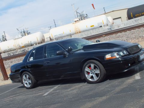 2003 Ford Crown Victoria - Side profile of a black sedan with chrome wheels and red brake calipers parked in front of rail tank cars and a warehouse.