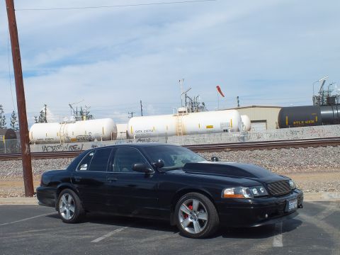 2003 Ford Crown Victoria - black sedan on chrome wheels with red brake calipers sits at an angle in a lot beside rail tank cars under a blue sky.