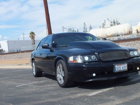 2003 Ford Crown Victoria - black sedan with a billet grille and halo headlights parked in a lot near rail tanks, wearing a California EVIL VIC plate.