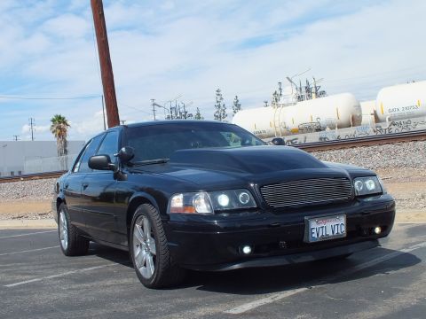 2003 Ford Crown Victoria - Front three-quarter view of a black sedan with chrome wheels, billet grille and an EVIL VIC California plate beside the railroad.