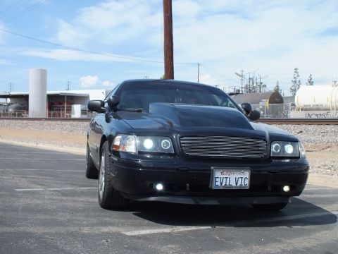 2003 Ford Crown Victoria - black sedan shown head-on with a billet grille, glowing halo headlights and an EVIL VIC plate, parked under a clear sky.