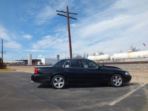 2003 Ford Crown Victoria - Side profile of a black sedan on chrome wheels parked in a lot beside railroad tracks and tank cars under a partly cloudy sky.