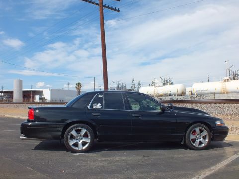 2003 Ford Crown Victoria - Side profile of a black sedan with chrome wheels and red brake calipers parked beside railroad tracks and industrial tanks.