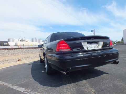 2003 Ford Crown Victoria - Rear three-quarter view of a black Ford Crown Victoria with Ford oval, SVT badge and an EVIL VIC plate, parked near rail tanks.