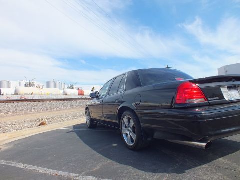2003 Ford Crown Victoria - Rear three-quarter view of a black sedan with a chrome wheel and dual exhaust parked beside railroad tracks and storage tanks.