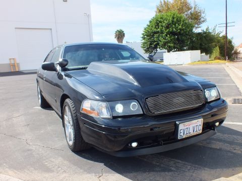 2003 Ford Crown Victoria - modified black full-size sedan with a cowl-induction hood, billet grille, and halo projector headlights sits in a parking lot.