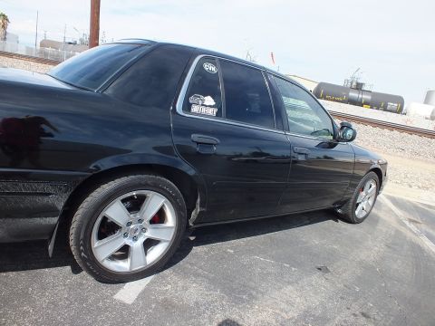 2003 Ford Crown Victoria - Rear quarter of a black sedan on chrome five-spoke wheels with window decals, parked near railroad tank cars.