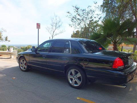 2003 Ford Crown Victoria - black full-size sedan with a trunk spoiler and chrome five-spoke wheels parked on a hilltop road by a palm tree.