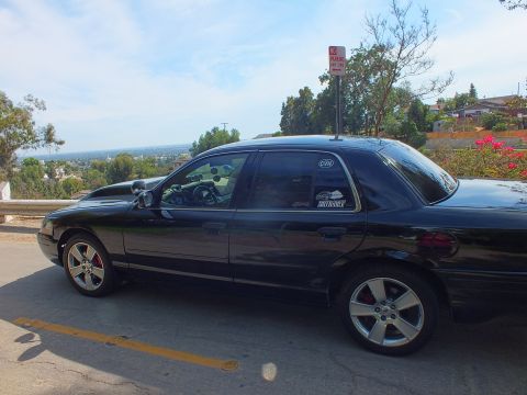 2003 Ford Crown Victoria - Passenger-side profile of a black full-size sedan with a cowl hood and chrome five-spoke wheels on an overlook road.