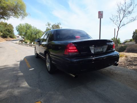 2003 Ford Crown Victoria - Rear three-quarter of a black full-size sedan with dual exhaust and dark taillights on a curving hill road.