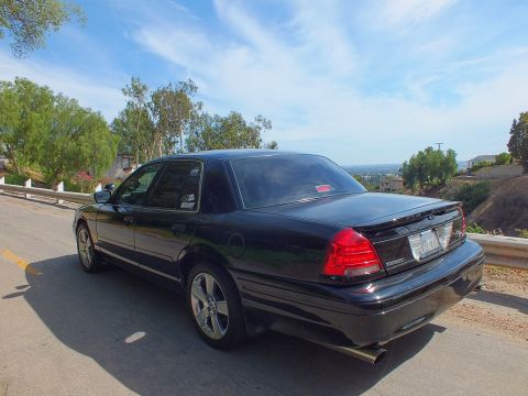 2003 Ford Crown Victoria - Rear driver-side view of a black full-size sedan with chrome wheels and a trunk spoiler overlooking a valley.