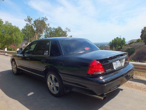 2003 Ford Crown Victoria - black full-size sedan with dual exhaust and red brake calipers shown rear three-quarter on a hill road.