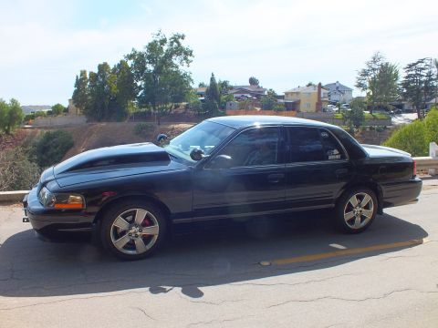 2003 Ford Crown Victoria - Driver-side profile of a black sedan with a cowl hood, red brake calipers, and chrome five-spoke wheels.