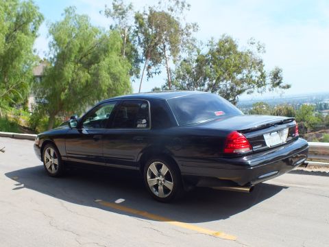 2003 Ford Crown Victoria - Rear driver-side view of a black full-size sedan with a trunk spoiler and dual exhaust on a hilltop road.