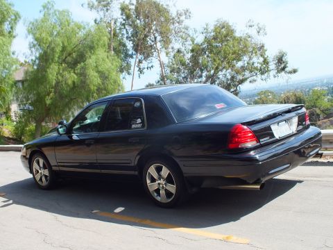2003 Ford Crown Victoria - Rear driver-side view of a black full-size sedan with dual exhaust and tinted windows on a tree-lined hill road.