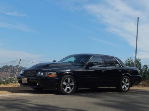 2003 Ford Crown Victoria - black full-size sedan with a billet grille, halo headlights, and a lowered stance parked under a blue sky.