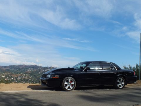2003 Ford Crown Victoria - black full-size sedan with a cowl hood and chrome wheels parked on a hilltop under a wide cloudy sky.