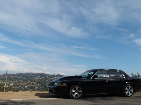 2003 Ford Crown Victoria - black full-size sedan parked at a hilltop overlook, framed small beneath a sweeping cloudy sky.