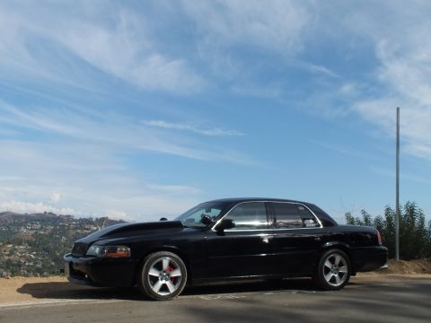 2003 Ford Crown Victoria - Driver-side profile of a black full-size sedan with chrome five-spoke wheels and red calipers on a hilltop.