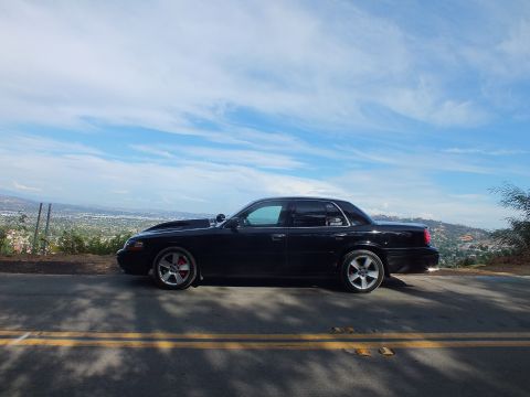 2003 Ford Crown Victoria - Driver-side profile of a lowered black full-size sedan parked on a sunlit road above a hazy valley.