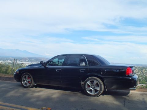 2003 Ford Crown Victoria - Rear driver-side view of a black full-size sedan with red calipers and chrome wheels, distant mountains beyond.