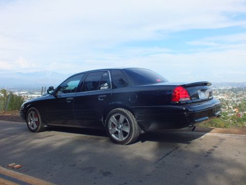 2003 Ford Crown Victoria - Rear three-quarter of a black full-size sedan with a trunk spoiler and chrome wheels above a mountain valley.