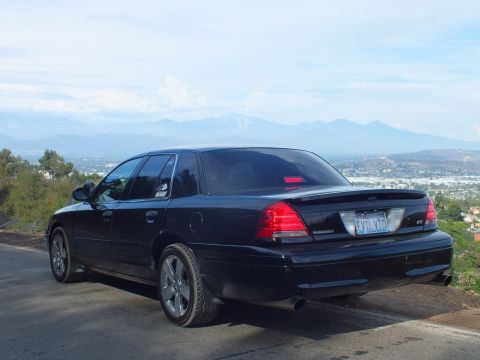 2003 Ford Crown Victoria - Rear three-quarter view of a black Ford Crown Victoria sedan on a scenic hilltop road, valley and mountains visible behind.