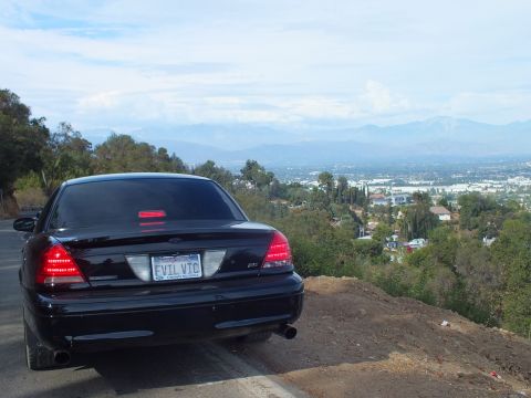 2003 Ford Crown Victoria - Rear three-quarter view of a black Ford Crown Victoria sedan parked at a dirt-edged ridge with a sprawling valley below.