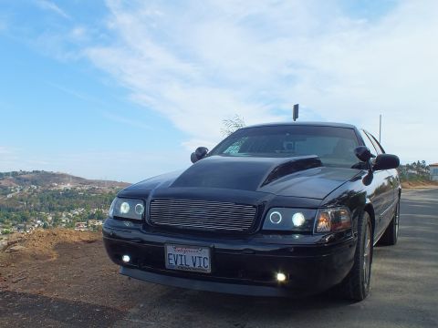 2003 Ford Crown Victoria - customized black Ford Crown Victoria seen from the front three-quarter, billet grille and halo headlights lit, parked at a dirt overlook.
