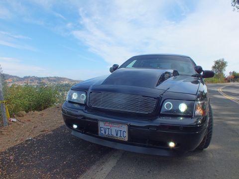 2003 Ford Crown Victoria - Low front view of a customized black Ford Crown Victoria with a chrome billet grille, glowing halo headlights, and an 'EVIL VIC' plate.
