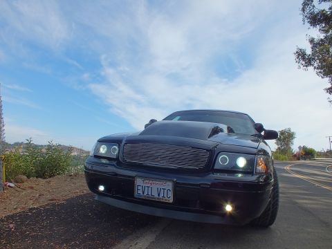 2003 Ford Crown Victoria - Front three-quarter view of a customized black Ford Crown Victoria with billet grille and halo headlights under a bright blue sky.