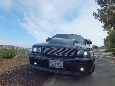 2003 Ford Crown Victoria - Low front view of a customized black Ford Crown Victoria, billet grille, halo headlights and fog lights lit, roadside under a blue sky.