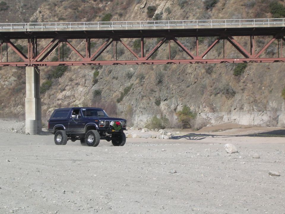 Dark lifted SUV parked on a dry riverbed gravel flat beneath a red steel truss bridge