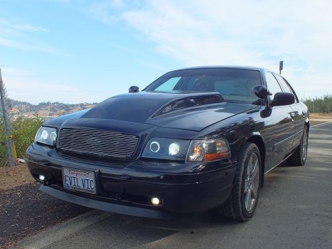 2003 Ford Crown Victoria - Front three-quarter view of a customized black Ford Crown Victoria with chrome billet grille, halo headlights, and 'EVIL VIC' plate.