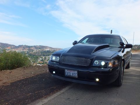 2003 Ford Crown Victoria - Front three-quarter view of a customized black Ford Crown Victoria with billet grille and halo headlights, hills in the background.
