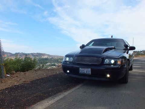 2003 Ford Crown Victoria - Front three-quarter view of a customized black Ford Crown Victoria with chrome billet grille and halo headlights parked on a roadside.