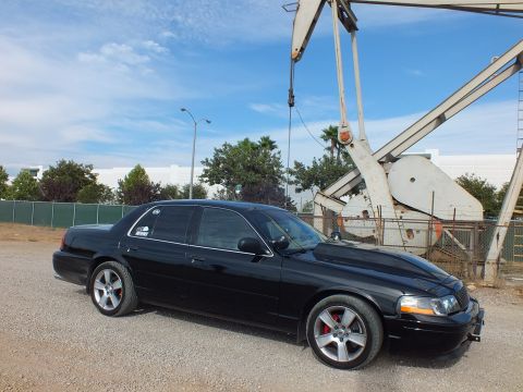 2003 Ford Crown Victoria - blacked-out full-size sedan with mesh grille and red brake calipers parked on gravel beside an oil pump jack.