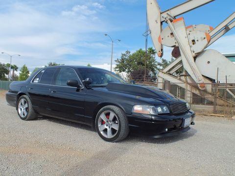 2003 Ford Crown Victoria - Front three-quarter view of a blacked-out full-size sedan with mesh grille and red brake calipers parked beside an oil pump jack.