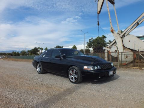 2003 Ford Crown Victoria - blacked-out full-size sedan with mesh grille and halo headlights parked on gravel near a tall oil pump jack.