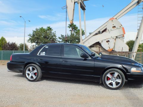 2003 Ford Crown Victoria - Side profile of a blacked-out full-size sedan with machined-spoke wheels and red brake calipers parked beside an oil pump jack.