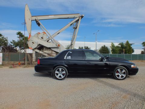 2003 Ford Crown Victoria - Side profile of a blacked-out full-size sedan parked on gravel in front of a large oil pump jack under a blue sky.