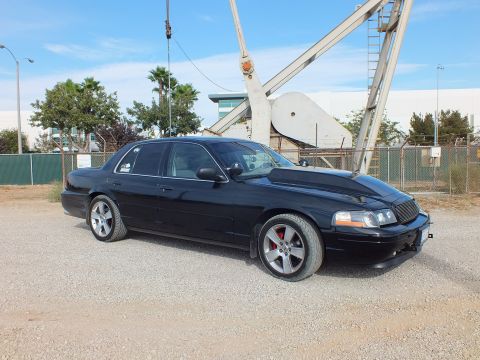 2003 Ford Crown Victoria - Front three-quarter view of a blacked-out full-size sedan with mesh grille and red brake calipers parked beneath an oil pump jack.