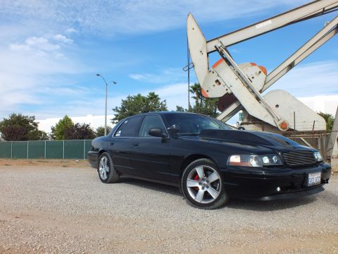 2003 Ford Crown Victoria - blacked-out full-size sedan with a chrome billet grille and machined wheels sits on gravel in front of a weathered oil pump jack.