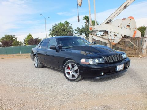 2003 Ford Crown Victoria - Front three-quarter shot of a glossy black sedan with billet grille and chrome wheels parked beside an industrial oil pump jack.