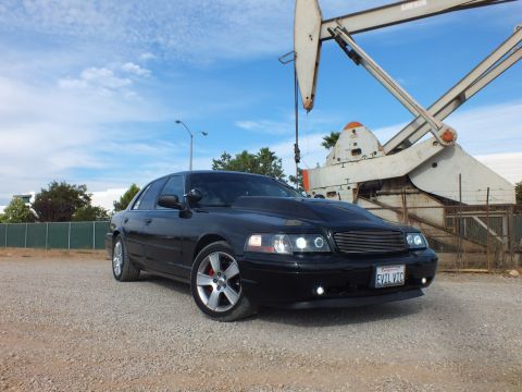 2003 Ford Crown Victoria - black sedan with halo headlights, billet grille and a California EVIL VIC plate sits on gravel before an oil pump jack.