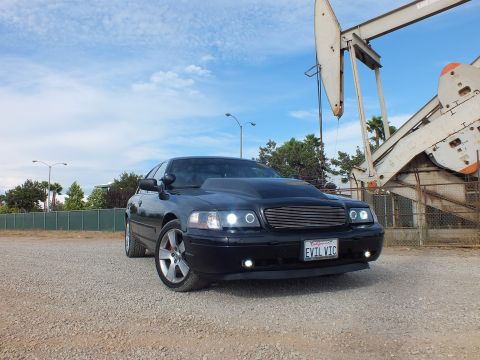2003 Ford Crown Victoria - Low-angle front view of a black sedan with glowing halo headlights and a California EVIL VIC license plate near an oil derrick.