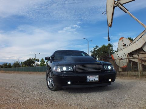 2003 Ford Crown Victoria - Head-on view of a black sedan with illuminated halo headlights, billet grille and EVIL VIC plate, an oil pump jack at right.
