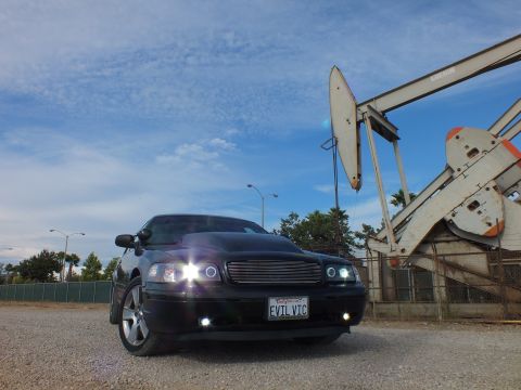 2003 Ford Crown Victoria - black sedan glows with halo headlights and fog lamps on gravel, framed by a tall industrial oil pump jack.